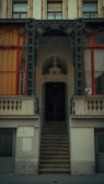An architectural facade featuring a slightly weathered staircase leading up to an ornate doorway. The building has decorative ironwork around the main entrance and two large windows on either side, framed with wooden trim and covered with curtains in warm tones. There are intricate embellishments and stonework throughout the facade.