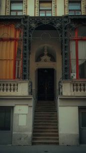 An architectural facade featuring a slightly weathered staircase leading up to an ornate doorway. The building has decorative ironwork around the main entrance and two large windows on either side, framed with wooden trim and covered with curtains in warm tones. There are intricate embellishments and stonework throughout the facade.