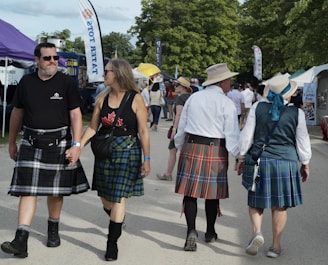 People walking at an outdoor event, with some wearing traditional kilts. Tents and banners are visible in the background with greenery surrounding the area. The scene has a relaxed and festive atmosphere.
