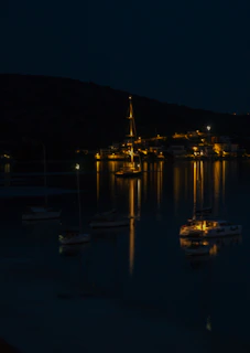 Evening scene of a sailboat anchored near a quiet shore with warm cabin lights glowing.