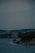 A coastal town with residential buildings and lush greenery lining the shoreline. Small boats are anchored in the calm water near the coast. The sky is overcast, creating a muted atmosphere.