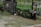 In a rural outdoor setting, several goats are scattered around, near a makeshift wooden shelter and a wooden pallet. A duck is present among the animals, next to two lying ducks. The environment is grassy with patches of dirt and surrounded by wooden fencing.