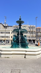 A teal-colored fountain adorned with sculptural griffins is situated in an urban square. Water streams from multiple levels of the fountain, and in the background, there are several multi-story buildings with a mix of architectural styles. Cars and a pedestrian are visible, and the sky above is clear and blue.