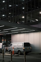 A silver van is parked in an underground parking area, partially illuminated by overhead lights. The background reveals a multi-level parking structure with other vehicles visible through metal framework and lighting. Orange traffic cones and barriers are positioned around the entry control area.