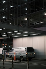 A silver van is parked in an underground parking area, partially illuminated by overhead lights. The background reveals a multi-level parking structure with other vehicles visible through metal framework and lighting. Orange traffic cones and barriers are positioned around the entry control area.