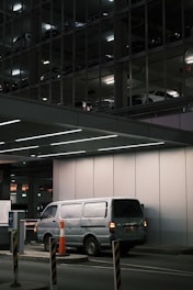 A silver van is parked in an underground parking area, partially illuminated by overhead lights. The background reveals a multi-level parking structure with other vehicles visible through metal framework and lighting. Orange traffic cones and barriers are positioned around the entry control area.