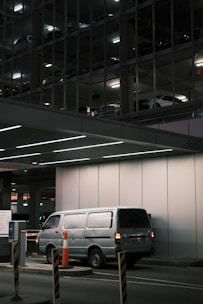 A silver van is parked in an underground parking area, partially illuminated by overhead lights. The background reveals a multi-level parking structure with other vehicles visible through metal framework and lighting. Orange traffic cones and barriers are positioned around the entry control area.