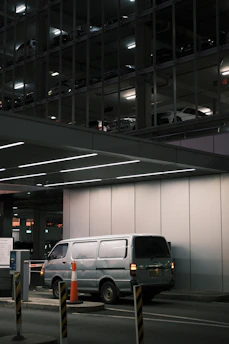A silver van is parked in an underground parking area, partially illuminated by overhead lights. The background reveals a multi-level parking structure with other vehicles visible through metal framework and lighting. Orange traffic cones and barriers are positioned around the entry control area.