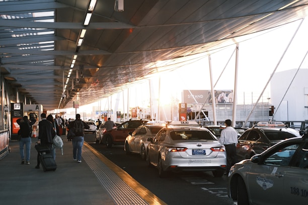 Several taxis are parked along the side of a road at an airport terminal. People with luggage walk towards and away from the taxis. The structure above is modern and metallic, casting shadows and reflecting the setting sun.