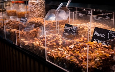 A variety of nuts and dried fruits are displayed in transparent containers at a market or store. Each section is labeled with its specific mix, and metal scoops are placed on the side for serving. The selection includes almonds, raisins, and other mixed nuts.