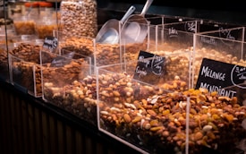 A variety of nuts and dried fruits are displayed in transparent containers at a market or store. Each section is labeled with its specific mix, and metal scoops are placed on the side for serving. The selection includes almonds, raisins, and other mixed nuts.