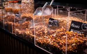 A variety of nuts and dried fruits are displayed in transparent containers at a market or store. Each section is labeled with its specific mix, and metal scoops are placed on the side for serving. The selection includes almonds, raisins, and other mixed nuts.