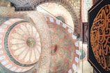 Intricate geometric and floral patterns adorn the domed ceiling of a traditional mosque, showcasing detailed Islamic art with vibrant colors. Arabic calligraphy is prominently featured against a richly decorated background.