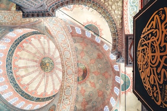 Intricate geometric and floral patterns adorn the domed ceiling of a traditional mosque, showcasing detailed Islamic art with vibrant colors. Arabic calligraphy is prominently featured against a richly decorated background.