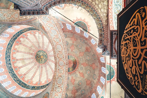 Intricate geometric and floral patterns adorn the domed ceiling of a traditional mosque, showcasing detailed Islamic art with vibrant colors. Arabic calligraphy is prominently featured against a richly decorated background.