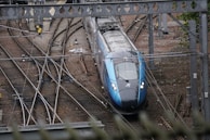 A modern train is navigating through a network of railway tracks, surrounded by various rail infrastructure elements, such as overhead wires and signal equipment. The scene includes multiple tracks converging and diverging, with the train prominently positioned in the center. The environment appears industrial with visible metal structures and gravel-covered ground.