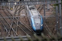 A modern train is navigating through a network of railway tracks, surrounded by various rail infrastructure elements, such as overhead wires and signal equipment. The scene includes multiple tracks converging and diverging, with the train prominently positioned in the center. The environment appears industrial with visible metal structures and gravel-covered ground.
