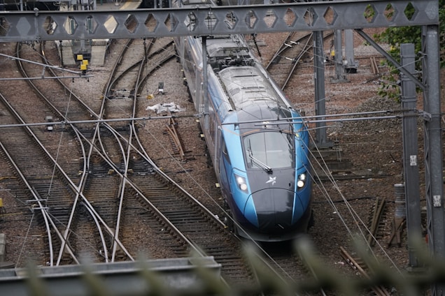 A modern train is navigating through a network of railway tracks, surrounded by various rail infrastructure elements, such as overhead wires and signal equipment. The scene includes multiple tracks converging and diverging, with the train prominently positioned in the center. The environment appears industrial with visible metal structures and gravel-covered ground.