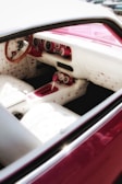 The interior of a car featuring a vintage design with plush white seats adorned with pink buttons. The dashboard and steering wheel are crafted from a mix of white and pink materials, complemented by an elegant wooden steering wheel. The overall design displays a retro aesthetic.
