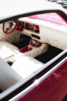The interior of a car featuring a vintage design with plush white seats adorned with pink buttons. The dashboard and steering wheel are crafted from a mix of white and pink materials, complemented by an elegant wooden steering wheel. The overall design displays a retro aesthetic.