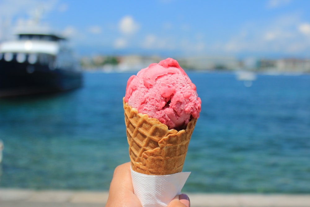 a hand holding an ice cream cone with a boat in the background