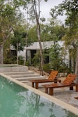 A serene outdoor scene featuring a rectangular swimming pool with light green water. Beside the pool, there are wooden lounge chairs placed on a concrete surface, along with steps leading up to a raised area that could potentially be a hot tub. The environment is surrounded by lush greenery and tall trees, with the presence of a modern, minimalist building in the background.