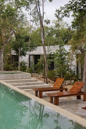 A serene outdoor scene featuring a rectangular swimming pool with light green water. Beside the pool, there are wooden lounge chairs placed on a concrete surface, along with steps leading up to a raised area that could potentially be a hot tub. The environment is surrounded by lush greenery and tall trees, with the presence of a modern, minimalist building in the background.