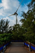 A large wind turbine stands prominently against a partly cloudy sky, surrounded by lush green trees. A wooden path bordered by fences and blue panels leads through the foliage, creating a harmonious blend of nature and technology.