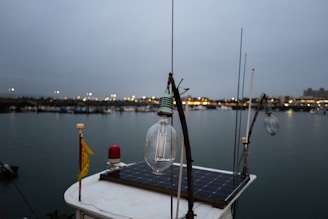 A close-up of a light bulb on a boat with a solar panel, set against a backdrop of a harbor. The evening sky is overcast and the distant lights from the shore reflect off the calm water. Fishing and other boats can be seen in the background.