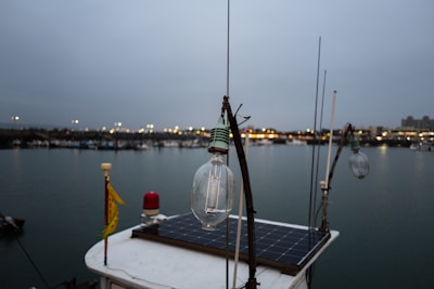 A close-up of a light bulb on a boat with a solar panel, set against a backdrop of a harbor. The evening sky is overcast and the distant lights from the shore reflect off the calm water. Fishing and other boats can be seen in the background.