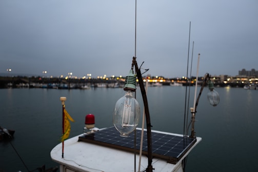A close-up of a light bulb on a boat with a solar panel, set against a backdrop of a harbor. The evening sky is overcast and the distant lights from the shore reflect off the calm water. Fishing and other boats can be seen in the background.