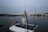 A close-up of a light bulb on a boat with a solar panel, set against a backdrop of a harbor. The evening sky is overcast and the distant lights from the shore reflect off the calm water. Fishing and other boats can be seen in the background.