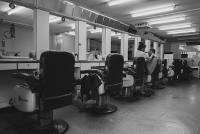 A barbershop interior with several empty reclining chairs lined up along a mirrored wall. A barber is seen attending to a customer, with various hairdressing tools and bottles organized along the countertop. The ambiance is quiet and orderly, with a tiled floor and fluorescent ceiling lights creating a clean look.