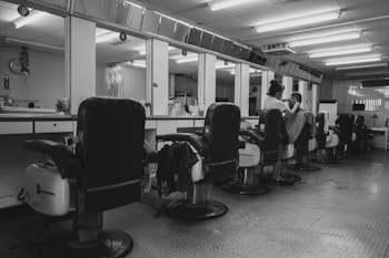 A barbershop interior with several empty reclining chairs lined up along a mirrored wall. A barber is seen attending to a customer, with various hairdressing tools and bottles organized along the countertop. The ambiance is quiet and orderly, with a tiled floor and fluorescent ceiling lights creating a clean look.