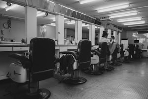 A barbershop interior with several empty reclining chairs lined up along a mirrored wall. A barber is seen attending to a customer, with various hairdressing tools and bottles organized along the countertop. The ambiance is quiet and orderly, with a tiled floor and fluorescent ceiling lights creating a clean look.