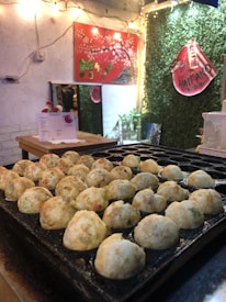 A close-up view of multiple round fried snacks on a griddle in a cozy indoor setting. The background includes a red artistic wall hanging with floral designs, a small table with some objects and a menu, as well as a green, plant-covered wall with a sign. Fairy lights add a warm and inviting ambiance to the scene.