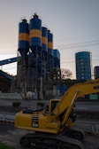 An industrial scene features a large yellow excavator in the foreground, labeled PC 210, with three tall cylindrical silos in the background. The silos are painted with alternating blue and orange bands. Various industrial structures and power lines are visible, with trees and the sky providing a backdrop.