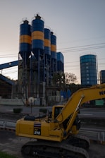 An industrial scene features a large yellow excavator in the foreground, labeled PC 210, with three tall cylindrical silos in the background. The silos are painted with alternating blue and orange bands. Various industrial structures and power lines are visible, with trees and the sky providing a backdrop.