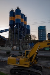 An industrial scene features a large yellow excavator in the foreground, labeled PC 210, with three tall cylindrical silos in the background. The silos are painted with alternating blue and orange bands. Various industrial structures and power lines are visible, with trees and the sky providing a backdrop.