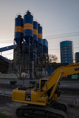 An industrial scene features a large yellow excavator in the foreground, labeled PC 210, with three tall cylindrical silos in the background. The silos are painted with alternating blue and orange bands. Various industrial structures and power lines are visible, with trees and the sky providing a backdrop.