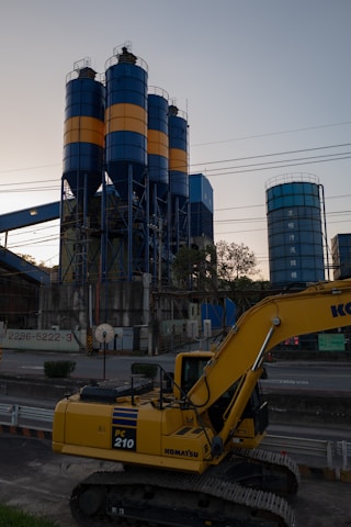 An industrial scene features a large yellow excavator in the foreground, labeled PC 210, with three tall cylindrical silos in the background. The silos are painted with alternating blue and orange bands. Various industrial structures and power lines are visible, with trees and the sky providing a backdrop.