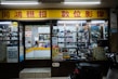 A storefront of a camera shop with various models of vintage cameras displayed on shelves behind the glass. The yellow and red signage features Chinese characters. Inside, the shop appears cluttered with boxes, film rolls, and photo equipment. There is a counter with a person sitting behind it, surrounded by more photographic items.
