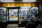 A storefront of a camera shop with various models of vintage cameras displayed on shelves behind the glass. The yellow and red signage features Chinese characters. Inside, the shop appears cluttered with boxes, film rolls, and photo equipment. There is a counter with a person sitting behind it, surrounded by more photographic items.