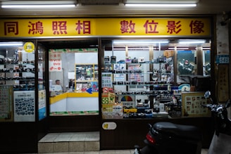 A storefront of a camera shop with various models of vintage cameras displayed on shelves behind the glass. The yellow and red signage features Chinese characters. Inside, the shop appears cluttered with boxes, film rolls, and photo equipment. There is a counter with a person sitting behind it, surrounded by more photographic items.