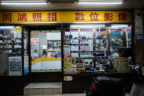 A storefront of a camera shop with various models of vintage cameras displayed on shelves behind the glass. The yellow and red signage features Chinese characters. Inside, the shop appears cluttered with boxes, film rolls, and photo equipment. There is a counter with a person sitting behind it, surrounded by more photographic items.