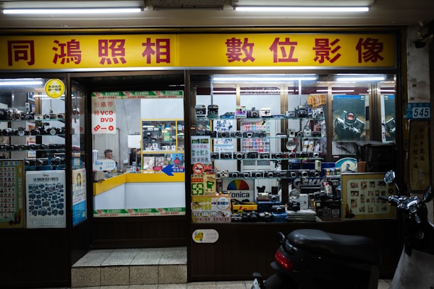 A storefront of a camera shop with various models of vintage cameras displayed on shelves behind the glass. The yellow and red signage features Chinese characters. Inside, the shop appears cluttered with boxes, film rolls, and photo equipment. There is a counter with a person sitting behind it, surrounded by more photographic items.