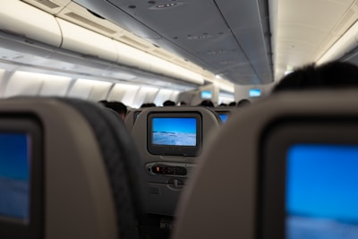 View of an airplane cabin from the perspective of a passenger. Several rows of seats with in-flight entertainment screens are visible. The cabin lights are on, and the image on the screen shows a landscape view, likely from an in-flight camera.