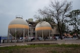 Two large, spherical industrial tanks are situated in an outdoor area, surrounded by a fence. These tanks are supported by multiple legs and feature a horizontal beige band around their circumference. In the background, there are trees with sparse foliage and a larger industrial building. The foreground consists of patches of grass and bare ground.