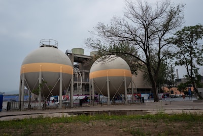 Two large, spherical industrial tanks are situated in an outdoor area, surrounded by a fence. These tanks are supported by multiple legs and feature a horizontal beige band around their circumference. In the background, there are trees with sparse foliage and a larger industrial building. The foreground consists of patches of grass and bare ground.