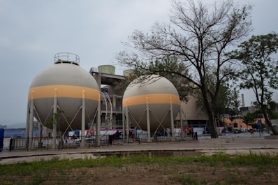 Two large, spherical industrial tanks are situated in an outdoor area, surrounded by a fence. These tanks are supported by multiple legs and feature a horizontal beige band around their circumference. In the background, there are trees with sparse foliage and a larger industrial building. The foreground consists of patches of grass and bare ground.